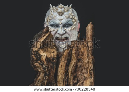 man or angry monster with thorns on face with futuristic makeup as alien with beard and lens in eyes on black background near tree
