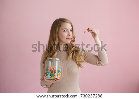 Isolated picture of playful charming young female looking at camera with sly expression and holding jelly bean, gummy bear or marmalade, taking it to her mouth as if teasing you. Candy, sweets