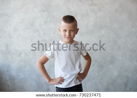 Studio picture of naughty mischievous Caucasian preschool boy dressed in casual white t-shirt holding hands on his waist and squinting eyes, planning another trick, showing his stubborn temper