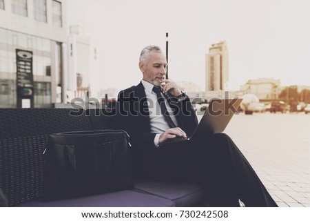 An old man with a beard in a strict business suit is sitting on a sofa in the street. On his lap is an open laptop. The old man works