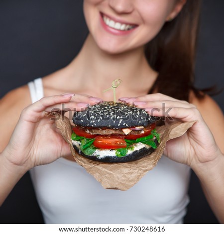 Close-up portrait of Young pretty woman with black burger in her hands on the dark grey background. Studio.