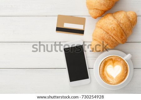Busy breakfast background. Hot foamy coffee and fresh croissants on wooden table with smartphone and credit card. Business person morning meals, top view, copy space