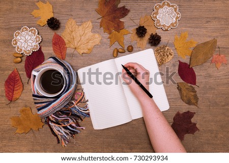 Writing woman hand on the blank space notebook and fall coffee in hands hugs on the wood table with dried maple leaves