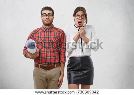 Worried female trainee has scared expression as looks at strict male teacher learns designing details, wants to improve knowledge. Frightened woman keeps hand together worry to have talk with boss