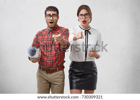 Miserable, discontent couple of creative workers, work with sketches and modern gadget, point at camera, asks someone else do work instead of them. Angry sorrorful man, woman gesture with fore fingers