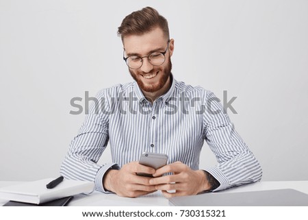 Happy fashionable male student sits on work place, prepares for classes, holds mobile phone, uses photo editing applications, posts new pictures via social networks, isolated over white background