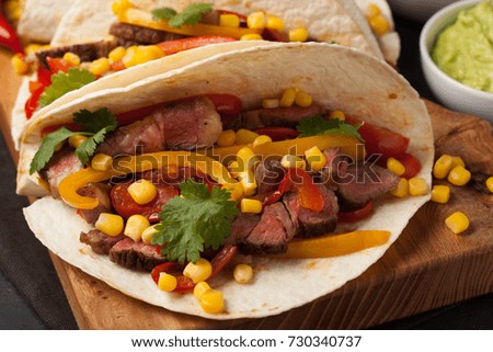 Three Mexican tacos with marbled beef, black Angus and vegetables on wooden Board on a dark stone background. Mexican dish with sauces guacamole and salsa in bowls.
