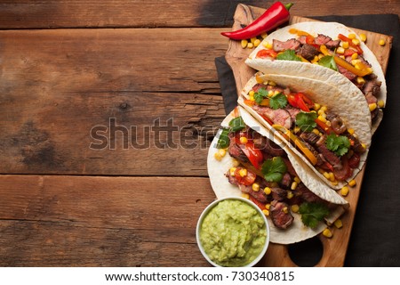 Three Mexican tacos with marbled beef, black Angus and vegetables on old rustic table. Mexican dish with sauces guacamole and salsa in bowls. Top view with copy space.