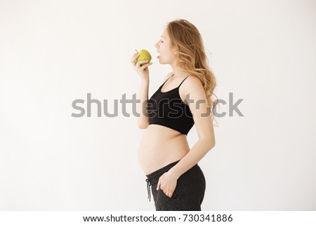 Portrait of good-looking young attractive pregnant mother with blond hair in home wear with open stomach, staying in profile, eating green apple, holding hand in pocket, living healthy life.