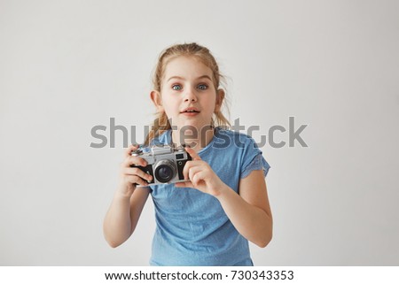 Portrait of good-looking blond girl in blue t-shirt holding camera in hands with concentrated expression, going to take a picture of cute cat on street.