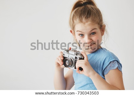 Portrait of funny little girl with blonde hair in tail hairstyle, looking in camera with silly expression, holding camera in hands going to take a picture.