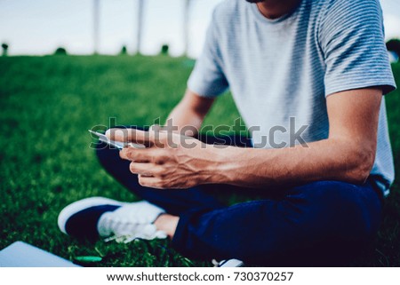Cropped image of young man in casual apparel sitting on green lawn during free time, hipster guy in stylish textile t-shirt recreating during weekends on grass near copy space for advertising