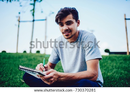 Portrait of talented male artist drawing sketch in notepad enjoying work outdoors, young  handsome hipster guy planning day making checklist and noting idea in personal diary sitting on green lawn