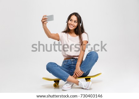 Portrait of young cheerful girl in white headphone makes selfie on smartphone, sitting on yellow skateboard, isolated over white background
