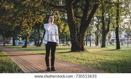 Nice and good picture of a young guy standing on the small road in a big green park and talking on the phone. The student decided to have a walk at the area after lessons in the evening.