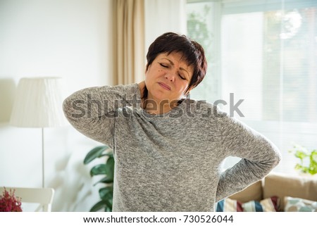 Mature woman suffering from backache at home. Massaging neck with hand, feeling exhausted, standing in living room.  Royalty-Free Stock Photo #730526044
