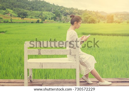 Asian woman in white dress using mobile phone and sitting on white wooden chair beside green rice field (vintage filter with flare yellow light)