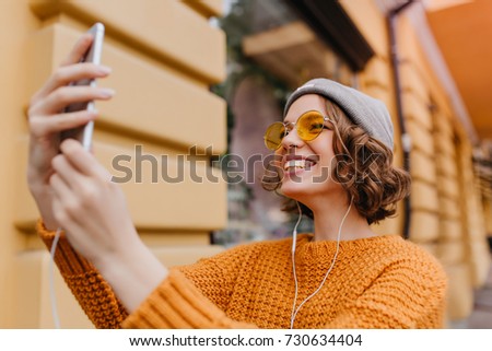 Lovely white girl with curly hairstyle making selfie in new outfit enjoying good weather. Outdoor portrait of ecstatic young woman in trendy sweater taking picture of herself beside old building.