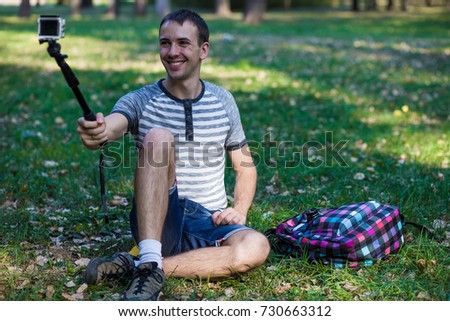 Young man with a backpack sitting on grass and taking selfie on an action camera