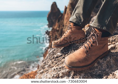 Man traveler sitting on mountain, closeup photo of hiking shoes. Boots on feet over sea landscape.