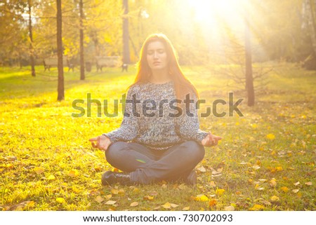 pretty young woman wearing warm fall sweater sitting alone on the ground in lotus meditation pose in the yellow autumn park
