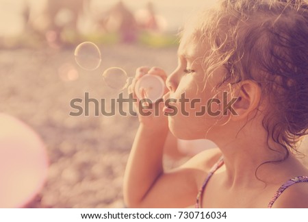 Five year old caucasian beautiful little girl blows soap bubbles on a summer beach on a sunny hot day. Happy carefree childhood. Toned orange purple effect. Royalty-Free Stock Photo #730716034
