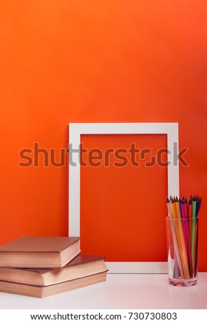 desk with white blank picture frame mock up. Pencils and books on orange background. Concept of teacher's day. Copy space