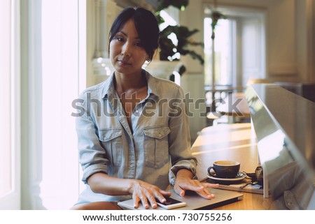 Young entrepreneur is working in a modern coworking space with a smartphone and a portable computer connected to wi-fi. Asian woman is looking at the camera while sitting with a laptop at the table.