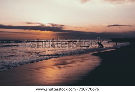 A silhouette of a surfer with a surfing board walking out the ocean on a dusk. Colorful sunset on a sandy beach of the tropical sea. Atmospheric photo of a nightfall on the ocean.