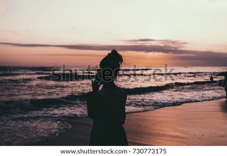 Young woman freelancer is taking photo of a beautiful and colorful sunset on the ocean beach by a smartphone. Photo of a silhouette of a hipster girl taking picture of a dusk sky and sea.