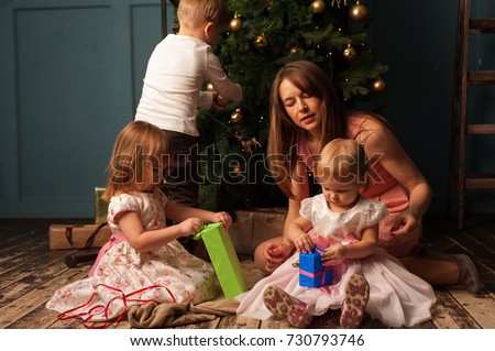 Loving mother and excited Caucasian children opening Christmas gifts next to tree. Happy parent and three kids having fun together.  
