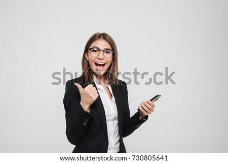 Portrait of a cheerful satisfied businesswoman in eyeglasses and suit holding mobile phone and showing thumbs up gesture isolated over white background
