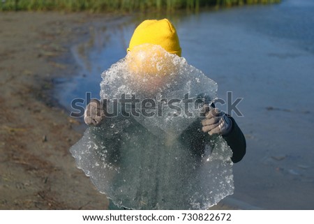 On the shore of the lake, the river is a child, the boy holds a plate of ice, hides, can not see the face. Winter clothes, jacket, yellow hat, gloves. First ice. Frost in autumn, winter background.