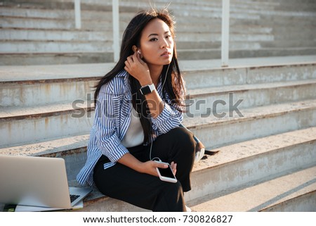 Photo of serious beautiful asian woman, listening to music, sitting on the stairs