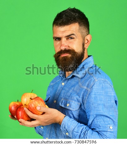 Farming and autumn products concept. Farmer with tricky smile and hands full of fresh apples. Guy presents homegrown harvest. Man with beard holds red fruit isolated on green background