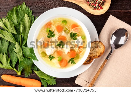 An overhead photo of a plate of chicken and noodles soup, shot from above on a dark rustic texture with a spoon, a wooden ladle with peppercorns, slices of bread, a celery branch, and a place for text
