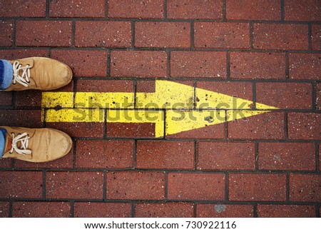 direction arrow on paving stones. man standing on paving slab and wear brown stylish leather shoes and blue jeans. yellow arrow. empty copy space for inscription