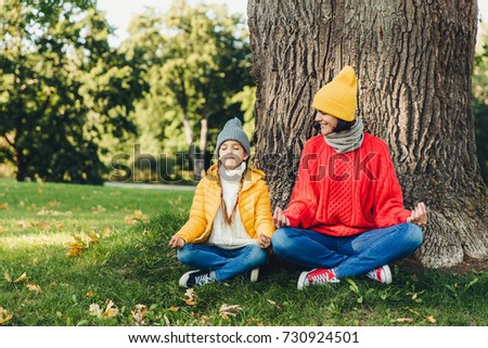 Body language concept. Carefree little kid sits near her beautiful mother, keep hands in mudra sign, keep eyes closed, try to calm down after hard work at home, admire fresh air in beautiful park