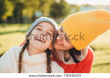 Beautiful female in yellow knitted hat kisses her adorable little child, have wonderful unforgettable time together. Small child smiles happily while receives kiss from mother. Family relationships