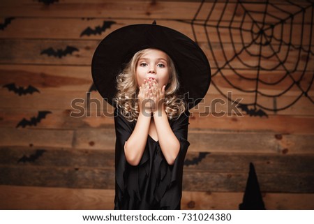 Halloween Witch concept - closeup shot of little caucasian witch child shocking face posing with bat and spider web on wooden studio background.