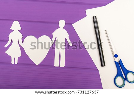 Paper silhouette of man and woman holding heart, scissors and marker near on a white sheet of paper on purple wooden background. Concept of family love