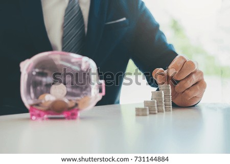 Businessman Inserting Coin In Transparent Piggybank Filled With Coins And House Model. Property Investment Concept