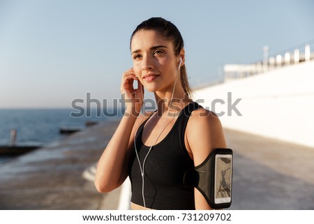 Picture of happy young sports woman listening music with earphones outdoors. Looking aside.