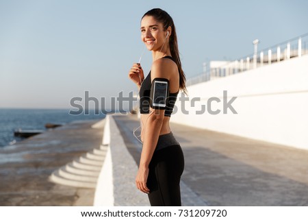 Picture of happy young sports woman listening music with earphones outdoors. Looking aside.