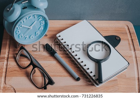 Alarm clock,spectacle,pen,magnifying glass and a note book on a wooden board with space for your text view from top.
