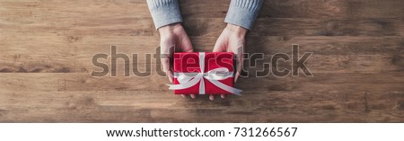 Woman in a gray sweater giving a christmas or birthday gift wrapped in red paper and white ribbon on a wooden table - top view, panoramic web banner with copy space