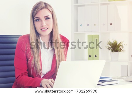 Beautiful young businesswoman wearing a red jacket is sitting in her office near a laptop. Toned image