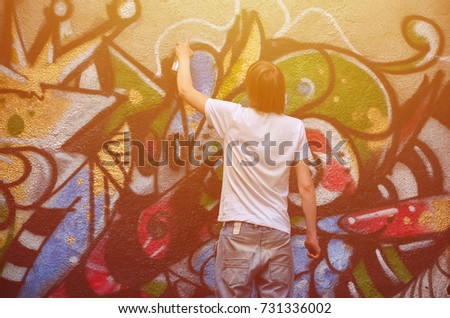 Photo in the process of drawing a graffiti pattern on an old concrete wall. Young long-haired blond guy draws an abstract drawing of different colors. Street art and vandalism concept