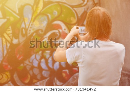 Photo in the process of drawing graffiti on an old concrete wall. A young, long-haired blond guy takes pictures of his completed drawing on a smartphone. Street art and vandalism concept