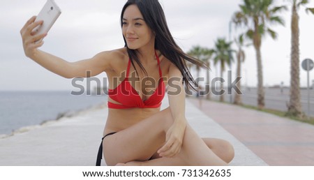 Excited young woman wearing bright bikini posing on seafront of tropical beach using smartphone and taking selfie smilingly.